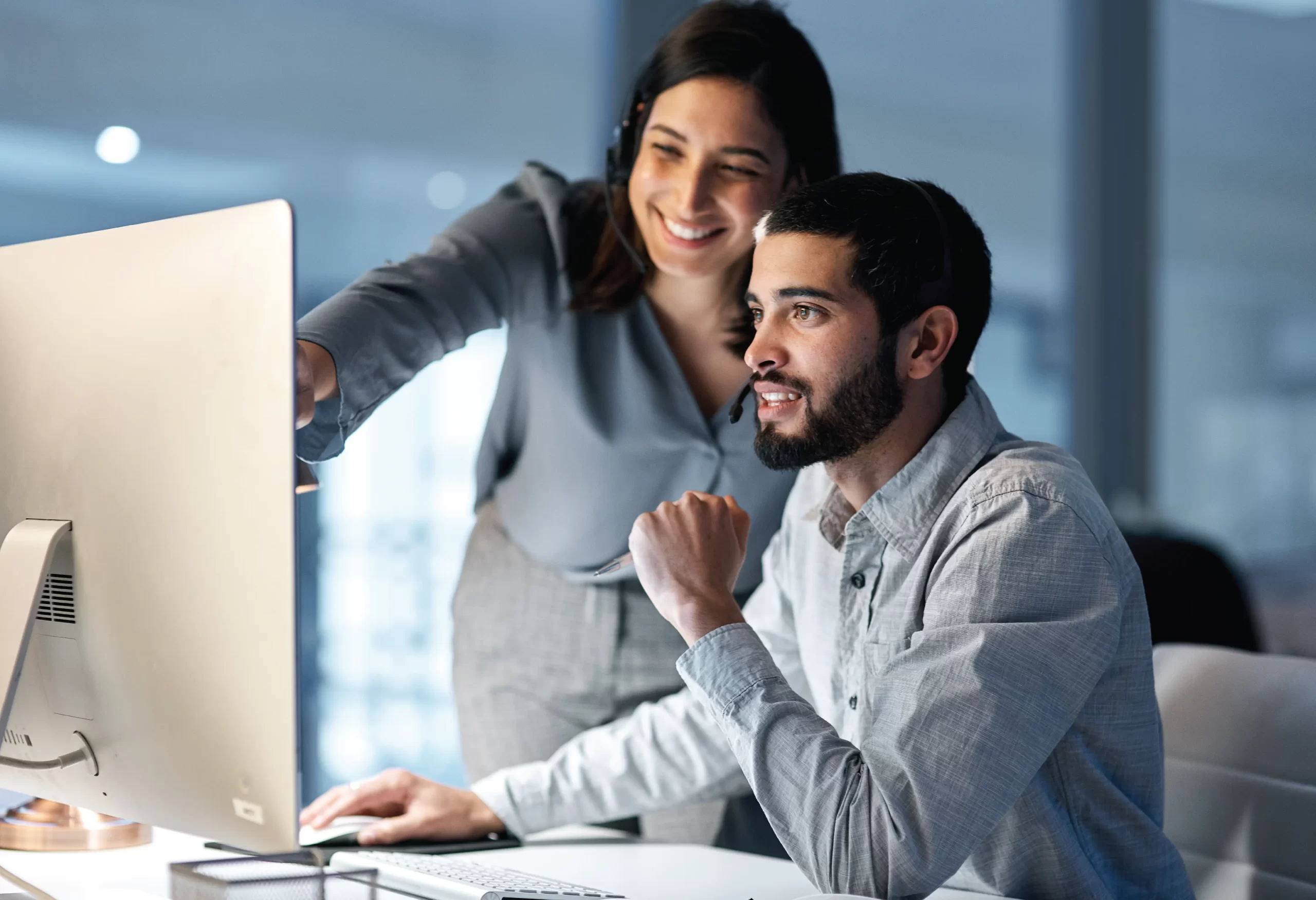 a man and woman sitting at a desk, with the woman pointing at the screen