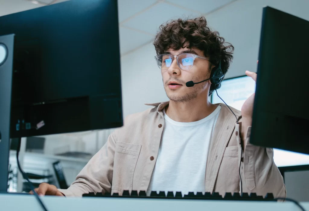 a young man sitting at two computer screens with a headset